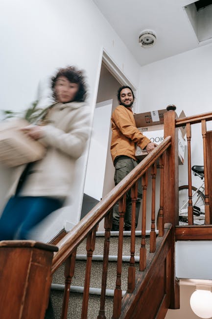 Inside a residential property during a house removal, a smiling man in a brown jacket is standing on a staircase landing, holding a cardboard box labeled 'cooking.' The staircase has polished wooden banisters and carpeted steps. A woman, slightly blurred to indicate movement, is walking past on the staircase, holding a package wrapped in plastic and cardboard, dressed in a light-colored coat. Behind the man, a doorway leads to another room, and part of a domestic indoor environment is visible, including a ceiling smoke detector and a bicycle partially seen behind the staircase. The scene captures the process of packing and moving items as part of home relocation, with packing materials and boxes stacked nearby, reflecting a professional yet casual approach by Man and a Van Brompton, specialists in removals and furniture transport service within the UK.