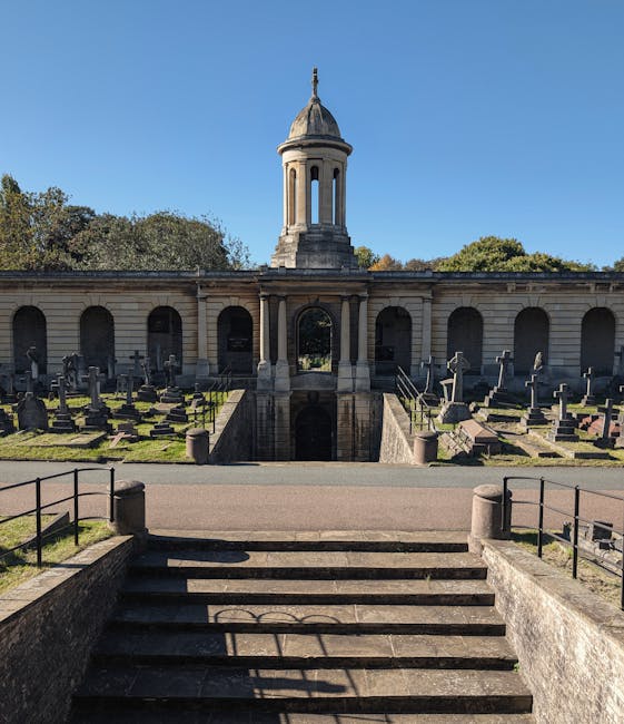 A wide-angle view of a historic crematorium building with a central domed tower and classical architectural details, situated behind a stepped stone staircase with metal railings on either side. The foreground shows the staircase leading up to the entrance area, with the steps casting shadows on the stone surface. Behind the staircase, there is a paved pathway and an outdoor space with grass and trees, leading to the crematorium's arched entrances, each flanked by tombstones and memorials. The building's facade features stonework and vertical window openings, set under a clear blue sky. Although the image is not directly related to house removals or moving services, it reflects the environment of a relocation process, with the potential for transport and careful handling of items in moving or packing contexts. The scene is well-lit with natural daylight, emphasizing the historic structure’s details. Man and a Van Brompton may utilize such sites for local relocations or transport logistics involving memorial or estate items, as part of their removals services.