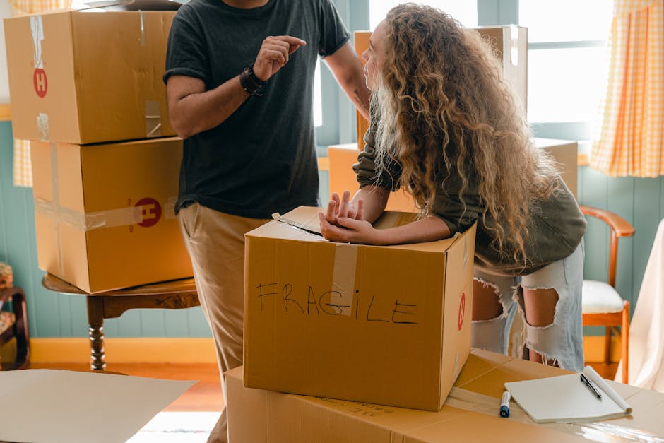 A person with light skin wearing beige trousers and a black T-shirt is standing inside a house, assisting a woman with light skin and curly blonde hair, who is leaning over a cardboard box labeled 'FR