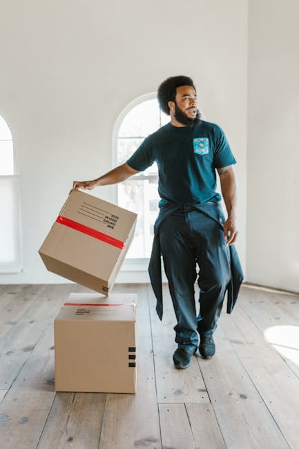 A person with light skin wearing beige trousers and a black T-shirt is standing inside a house, assisting a woman with light skin and curly blonde hair, who is leaning over a cardboard box labeled 'FR
