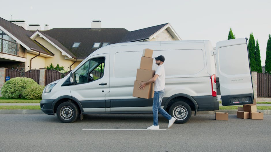 A man wearing a white shirt, blue jeans, white sneakers, and a black cap is involved in a home relocation process, carrying three cardboard boxes stacked in his arms and walking alongside a white panel van parked on a residential street. The van's side door is open for loading or unloading, with additional boxes placed on the pavement nearby. Inside a suburb with modern housing, the background features houses with pitched roofs, dormer windows, and small gardens with shrubs and trees. The scene is illuminated by daylight, indicating clear weather, and the man is engaged in the process of packing or unpacking for a furniture transport or moving service, with the van positioned adjacent to a driveway or curb for efficient loading as part of a professional removals service by Man and a Van Brompton.