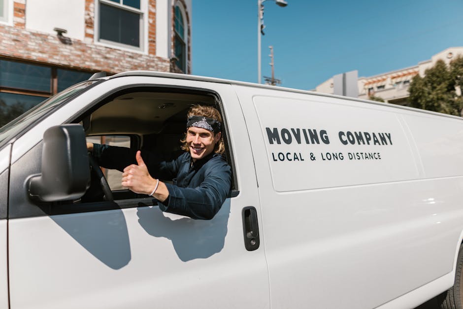 A young male mover with long hair, wearing a bandana and a dark long-sleeved shirt, sits in the driver's seat of a white van labeled with 'MOVING COMPANY' and 'LOCAL & LONG DISTANCE' on the side. He is smiling and giving a thumbs-up gesture towards the camera. The van is parked outside a residential building with a brick facade and large windows, with clear blue skies overhead. In the background, part of the building's exterior, some greenery, and utility poles are visible. The scene depicts a professional home relocation or furniture transport scenario, with the van positioned on the pavement near the property's entrance, ready for loading or unloading belongings during a house move facilitated by Man and a Van Brompton.