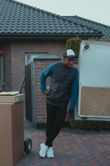 A man wearing a white shirt, blue jeans, white sneakers, and a black cap is involved in a home relocation process, carrying three cardboard boxes stacked in his arms and walking alongside a white panel van parked on a residential street. The van's side door is open for loading or unloading, with additional boxes placed on the pavement nearby. Inside a suburb with modern housing, the background features houses with pitched roofs, dormer windows, and small gardens with shrubs and trees. The scene is illuminated by daylight, indicating clear weather, and the man is engaged in the process of packing or unpacking for a furniture transport or moving service, with the van positioned adjacent to a driveway or curb for efficient loading as part of a professional removals service by Man and a Van Brompton.