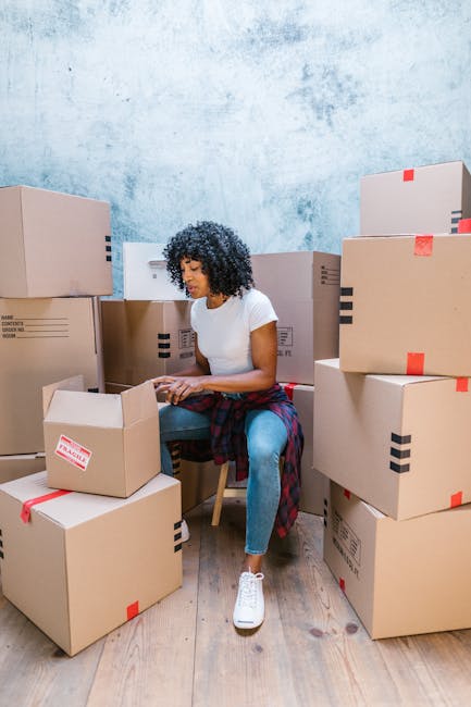 A young woman with long brown hair, sitting on the floor inside a home surrounded by numerous cardboard moving boxes, some sealed with red packing tape and others open, revealing packing materials. Sh